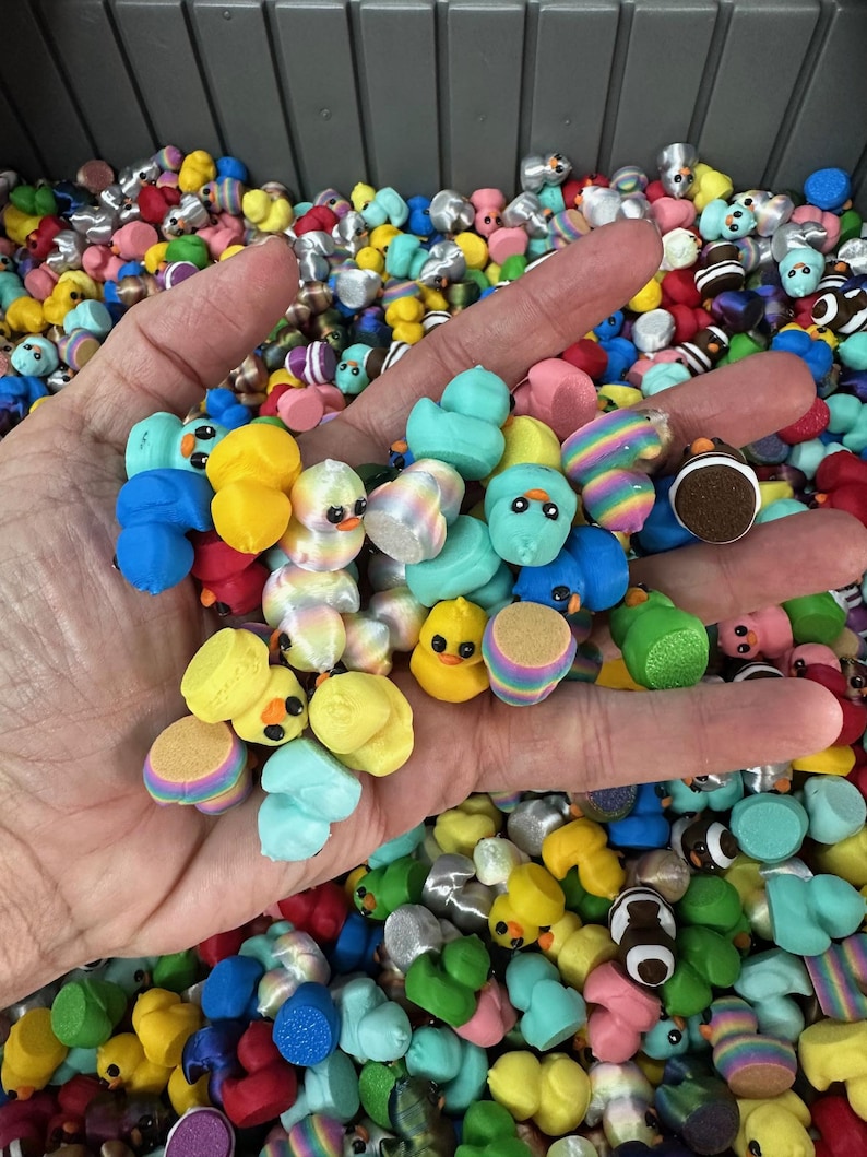May include: A hand holding a collection of colorful miniature duck figurines. The ducks are various colors, including yellow, blue, green, and pink, with some featuring striped or patterned designs. The background is filled with more of the same figurines.