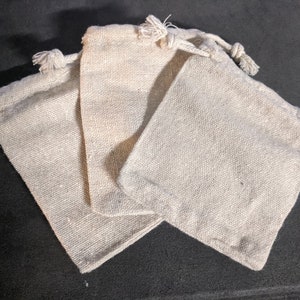 Puede incluir: Tres pequeñas bolsas de tela de color blanquecino con cordón de ajuste, hechas de un material natural similar al lino.