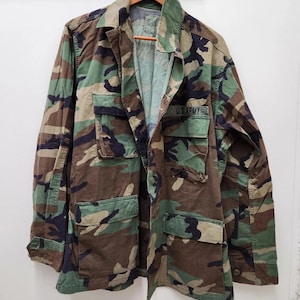May include: A camouflage pattern jacket with four front pockets and the text "U.S. ARMY" on the right chest. The jacket is made of a durable fabric and features a button-up front and a collared neckline. The colour palette includes shades of green, brown, and black.