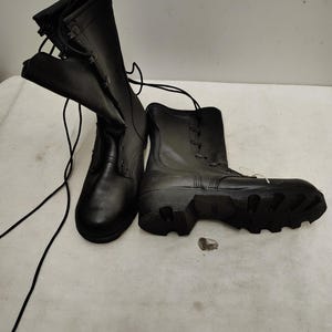 May include: A pair of black tactical combat boots featuring a high-top design, smooth leather construction, and thick rubber lug soles. The boots include long black laces and a reinforced heel area, with one boot standing upright and the other lying on its side.