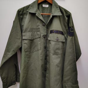 May include: Olive green US Air Force shirt with two chest pockets, button closures, and a collar. The shirt features the text "US AIR FORCE" and a patch on the sleeve. The shirt is hanging on a wooden hanger.