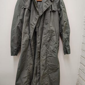 May include: An olive green trench coat with a button closure, shoulder straps, and a wide collar. The coat is displayed on a wooden hanger. The coat is made of a lightweight material and is hanging against a white wall.