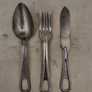 May include: A set of three vintage silver-colored utensils: a spoon, a fork, and a knife. Each piece has a handle with a teardrop-shaped opening and the letters "U.S." stamped on it. The spoon has a large, oval bowl.