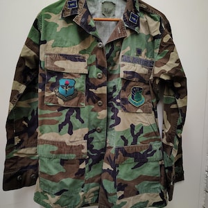 May include: A long-sleeved camouflage jacket featuring a woodland pattern in shades of green, brown, and black. The jacket includes a button-down front, four front pockets, collar insignia, and two embroidered patches with the text U.S. AIR FORCE.