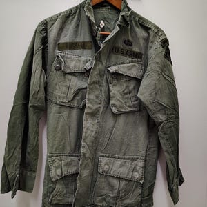 May include: An olive green military-style field jacket featuring a button-up front, four large front pockets with button flaps, and a high collar. The jacket displays patches with the text RUSSELL and U.S. ARMY, along with a shoulder insignia patch.
