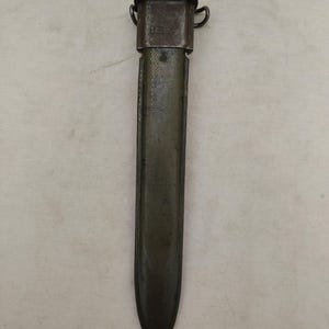 Op de afbeelding: Een olijfgroene metalen bajonetschede met een donkergrijze stalen monding en twee zijdelingse lussen. Het lichaam van de schede heeft een getextureerd oppervlak met zichtbare gebruikssporen, waaronder kleine krassen en verkleuringen.