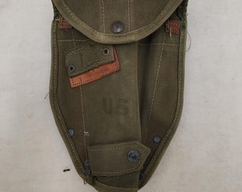 May include: An olive drab canvas shovel carrier featuring a fold-over flap with a metal snap closure, a small rectangular leather reinforcement patch with metal eyelets, and the letters US stamped in the center of the front panel.