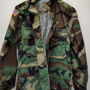 Peut inclure: Veste de camouflage avec plusieurs poches et le mot "ARMY" sur un écusson. Un écusson du drapeau américain est visible sur l'épaule gauche. La veste est suspendue à un cintre en bois.