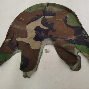 Puede incluir: Una funda de camuflaje para casco con un patrón de bosque de formas marrones, negras y verdes. La pieza de tela tiene una forma semicircular curva con bordes cosidos y varias aberturas pequeñas reforzadas a lo largo de la parte superior.