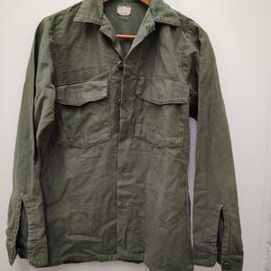 Puede incluir: Una camisa de manga larga abotonada en color verde oliva con cuello en punta, dos bolsillos en el pecho con solapas abotonadas y puños con botones. La camisa está confeccionada en tejido texturizado y tiene una etiqueta rectangular en el cuello.