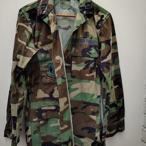 Peut inclure: Veste de camouflage avec plusieurs poches et l'inscription "U.S. AIR FORCE" sur le devant. La veste est un mélange de couleurs vertes, marron et noires. Elle est suspendue à un cintre en bois.
