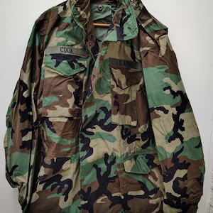 May include: A camouflage field jacket featuring a woodland pattern of green, brown, and black. The jacket has a high collar, long sleeves, and two chest pockets with flaps. Above the pockets, the text COOK and U.S. ARMY are embroidered.