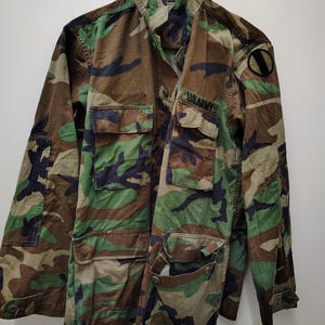 Puede incluir: Chaqueta militar con estampado de camuflaje con múltiples bolsillos y el texto "US ARMY". La chaqueta presenta una combinación de colores verde, marrón y negro. Se ve un parche en la parte superior del brazo.
