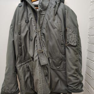 May include: A vintage olive green parka with a hood, button closures, and multiple pockets. The jacket is hanging on a wooden hanger. The parka has a worn appearance, suggesting it is a used or vintage item.
