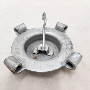 May include: A silver-colored metal ashtray with a circular design. It features four rounded protrusions and a central hook for hanging. The ashtray has a slightly worn appearance.