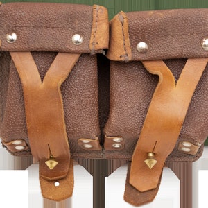 Könnte beinhalten: Eine braune Ledertasche mit zwei Fächern, mit hellbraunen Lederriemen und silbernen Nieten. Die Tasche hat eine strukturierte Oberfläche und eine Vintage-Ästhetik, mit einem kleinen messingfarbenen Stift an jedem Riemen. Vintage-Accessoire.
