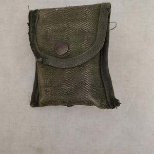 May include: A small, rectangular olive green canvas pouch featuring a fold-over flap secured with a single dark metal snap button. The edges of the pouch show visible fraying and wear along the stitched seams of the heavy-duty fabric.