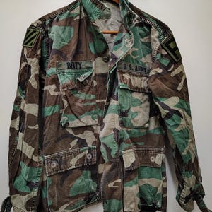 May include: A camouflage military jacket with multiple pockets and the text "BEITY" and "U.S. ARMY" on the front. The jacket features a green, brown, and tan pattern, with a button-up front and a collar. The jacket is hanging on a wooden hanger.