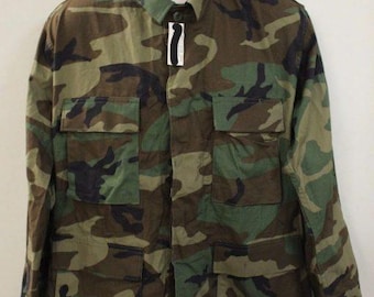 May include: A long-sleeve button-up shirt featuring a woodland camouflage pattern in shades of green, brown, and black. The shirt includes two chest pockets with flaps and two lower pockets with flaps, a standard collar, and a front button closure.