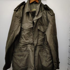 May include: An olive green field jacket featuring a collared neckline, long sleeves with buttoned cuffs, shoulder epaulets, and four front pockets with buttoned flaps. The jacket includes a central zipper closure and a drawstring waist detail.