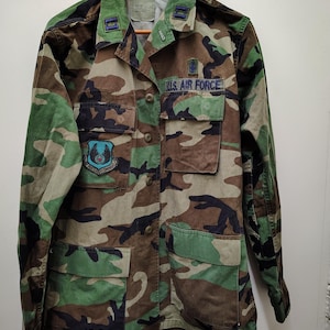 Puede incluir: Chaqueta de camuflaje con el texto "U.S. AIR FORCE" y parches. La chaqueta tiene múltiples bolsillos y una parte delantera abotonada. La combinación de colores incluye tonos de verde, marrón y negro.