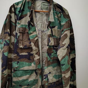 May include: A camouflage jacket in shades of green, brown, and black, featuring multiple pockets and a button-down front. The jacket is displayed on a wooden hanger, showcasing a military-style design.