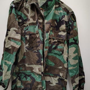 Peut inclure: Veste de camouflage aux motifs verts, marron et noirs. Elle est dotée de multiples poches, d'un col et de boutons. La veste est suspendue à un cintre en bois.