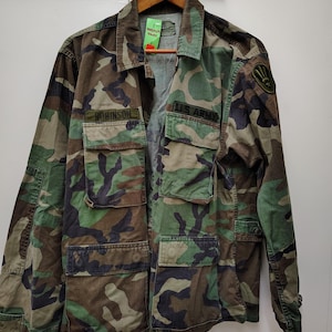 May include: A camouflage pattern field jacket featuring a collar, long sleeves, and four front pockets. The jacket displays a black embroidered name tag reading ROBINSON, a black U.S. ARMY patch, and a circular shoulder patch with a three-pronged emblem.