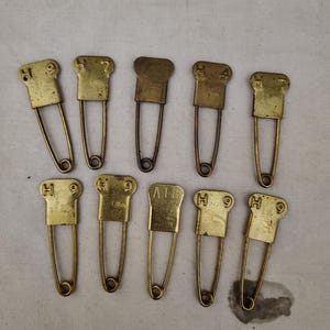 May include: A collection of ten vintage brass safety pins. Each pin features a rectangular head with stamped markings, including the letter "H" and numbers. The pins have a classic design with a curved wire and a clasp.