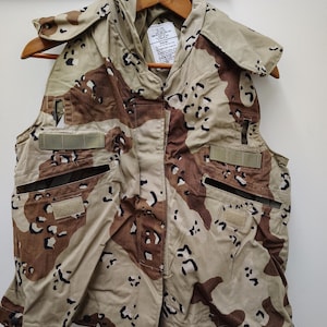 Puede incluir: Un chaleco de camuflaje desértico en tonos beige y marrón, con cuello alto y varios bolsillos. El chaleco está colgado de una percha de madera. El patrón es un diseño de camuflaje desértico.
