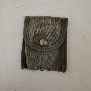 Puede incluir: Pequeño estuche de lona verde oliva con solapa redondeada y cierre de botón metálico. El estuche tiene un aspecto texturizado y un ribete verde oscuro. El fondo es de color claro y neutro.