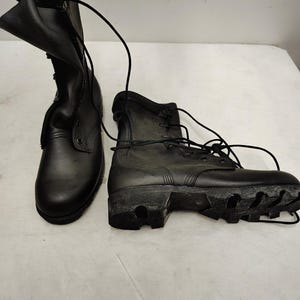 May include: A pair of black leather combat boots featuring a high-top design, rounded toes, and black laces. One boot stands upright while the other lies on its side, revealing a thick, textured black rubber lug sole with deep tread patterns.