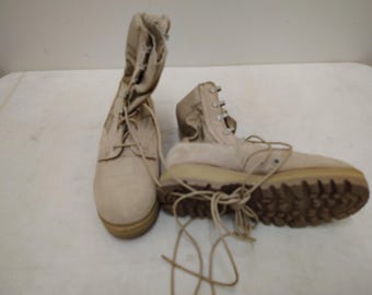 Op de afbeelding: Een paar beige suède laarzen met hoge schacht, voorzien van metalen oogjes voor de veters, lange beige veters en dikke, getextureerde bruine rubberen zolen. De ene laars staat rechtop terwijl de andere op zijn zijkant ligt.