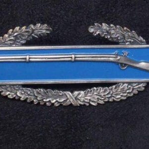 Può includere: Un distintivo militare argentato con sfondo smaltato blu. Il distintivo presenta un moschetto con canna bianca e una ghirlanda di foglie su entrambi i lati.