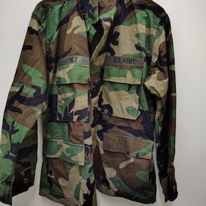 Puede incluir: Chaqueta de camuflaje con los textos "ARNEY" y "U.S.ARMY" en la parte delantera. La chaqueta tiene múltiples bolsillos y cierre de botones. La combinación de colores incluye tonos de verde, marrón y negro.