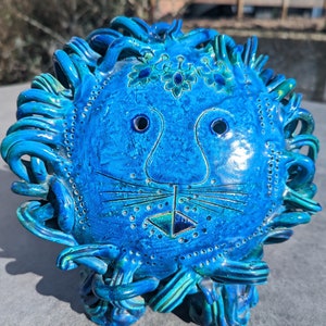 May include: A striking ceramic lion sculpture in shades of blue and turquoise. The lion's face has etched details, circular eye sockets, and a textured mane. This decorative art piece is a unique home decor item.
