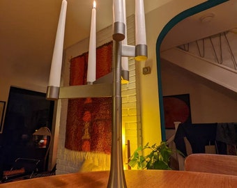 May include: A silver-toned candelabra with five lit white candles. The candelabra has a modern design with a flared base and horizontal arms holding the candles. The candles are tall and slender, with flames at the top. The candelabra is on a wooden table.