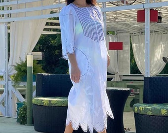 May include: A woman wearing sunglasses and a white, lace-trimmed maxi dress stands by a swimming pool. The pool is surrounded by white patio furniture and lush greenery.