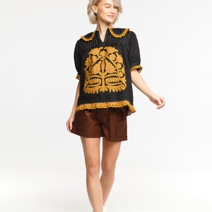 May include: A woman wearing a black linen blouse with a gold embroidered floral design and brown shorts. She is wearing white sneakers and has short blonde hair.