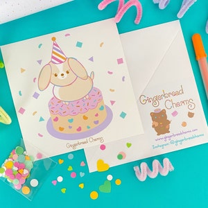 May include: A white card with a cartoon illustration of a dog wearing a party hat and sitting on a pink and purple cake with sprinkles. The card has the text "Gingerbread Charms" at the bottom. The card is on a blue background with confetti and other decorations.