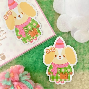 May include: A sticker of a cartoon dog wearing a green and white plaid coat, a pink scarf, and a pink hat with a bow. The dog is holding a small brown bag with a reindeer on it.