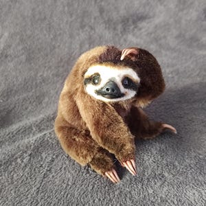 Puede incluir: Un juguete de peluche de perezoso hecho a mano con pelaje marrón, una cara blanca y marcas negras alrededor de los ojos. El perezoso está sentado con las garras visibles y su mano derecha tocando su cabeza. El juguete está sobre una superficie gris.