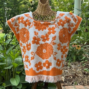 May include: A white terry cloth top featuring a vibrant orange floral pattern across the front and shoulders. The garment has a wide neckline, short sleeves, and a straight hemline finished with a fringe trim along the bottom edge.