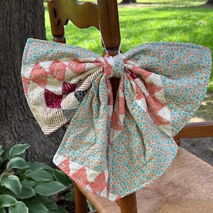 May include: A large decorative bow crafted from quilted fabric featuring a patchwork design. The fabric displays a mix of teal floral patterns, cream-colored sections, and coral-pink geometric shapes. The bow is tied with a central fabric knot.