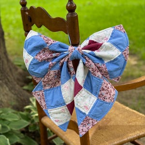May include: A large decorative bow constructed from quilted fabric patches in shades of blue, cream, and dusty rose with floral and vine patterns. The bow features a central knot and two long, pointed tails, showcasing a patchwork design.