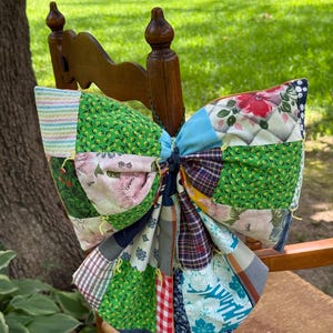 May include: A large decorative bow constructed from various fabric patches in a patchwork style. The bow features a mix of green floral prints, red and white gingham, blue plaid, and white fabric with pink floral patterns, tied with a dark center.