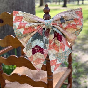 May include: A large, decorative bow made from a patchwork quilt, featuring various fabric patterns in shades of peach, cream, green, and burgundy. The bow is attached to the back of a wooden chair, creating a rustic, vintage aesthetic.