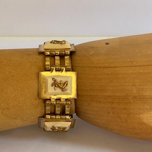 Puede incluir: Un brazalete de tono dorado con un diseño de cadena de eslabones y acentos cuadrados. Cada acento presenta un camafeo blanco con un diseño dorado.