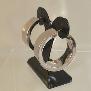 Peut inclure: Une paire de boucles d'oreilles créoles argentées sur un présentoir noir. Les boucles d'oreilles sont épaisses et de forme arrondie.