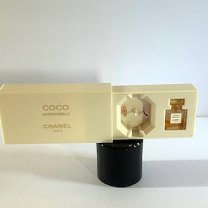 Könnte beinhalten: Eine weiße Box mit dem Text "COCO MADEMOISELLE CHANEL PARIS" auf der Oberseite. Die Box ist geöffnet und eine kleine durchsichtige Parfümflasche mit dem Text "COCO MADEMOISELLE CHANEL PARIS" ist im Inneren sichtbar.